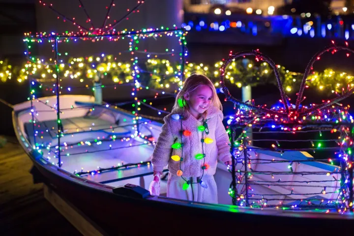 Child in a boat decorated with colorful holiday lights, smiling amidst festive glow.