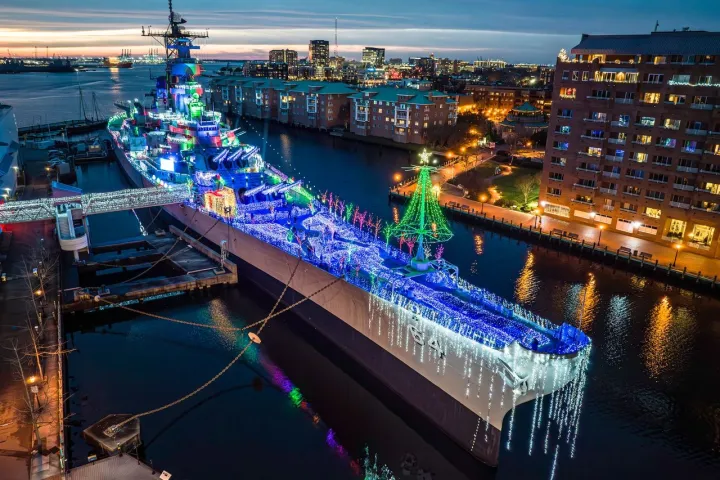 Battleship decorated with colorful Christmas lights docked in an urban harbor at dusk.