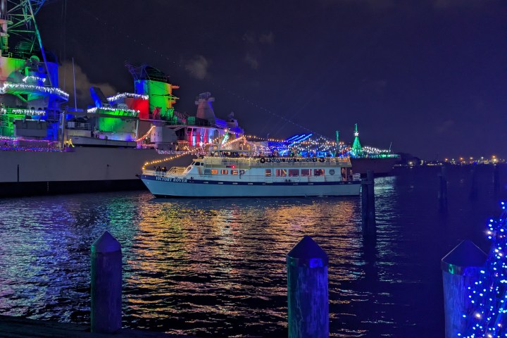 Boat with festive lights on water at night, near illuminated battleship.