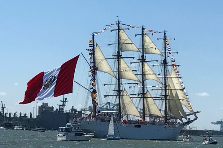 Sailing ship with large red and white flag in harbor, surrounded by smaller boats.